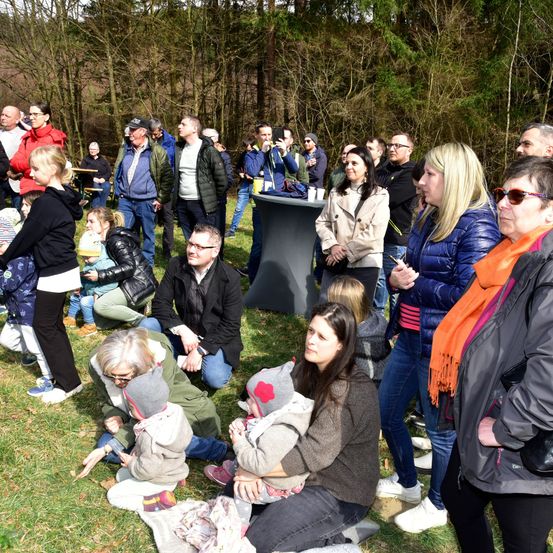 Eine große Gruppe von Menschen, darunter Erwachsene und Kinder, versammelt sich auf einem Grasfeld, wahrscheinlich für eine Gemeinschaftsveranstaltung. Einige knien, andere stehen und einige halten ihre Kinder.
