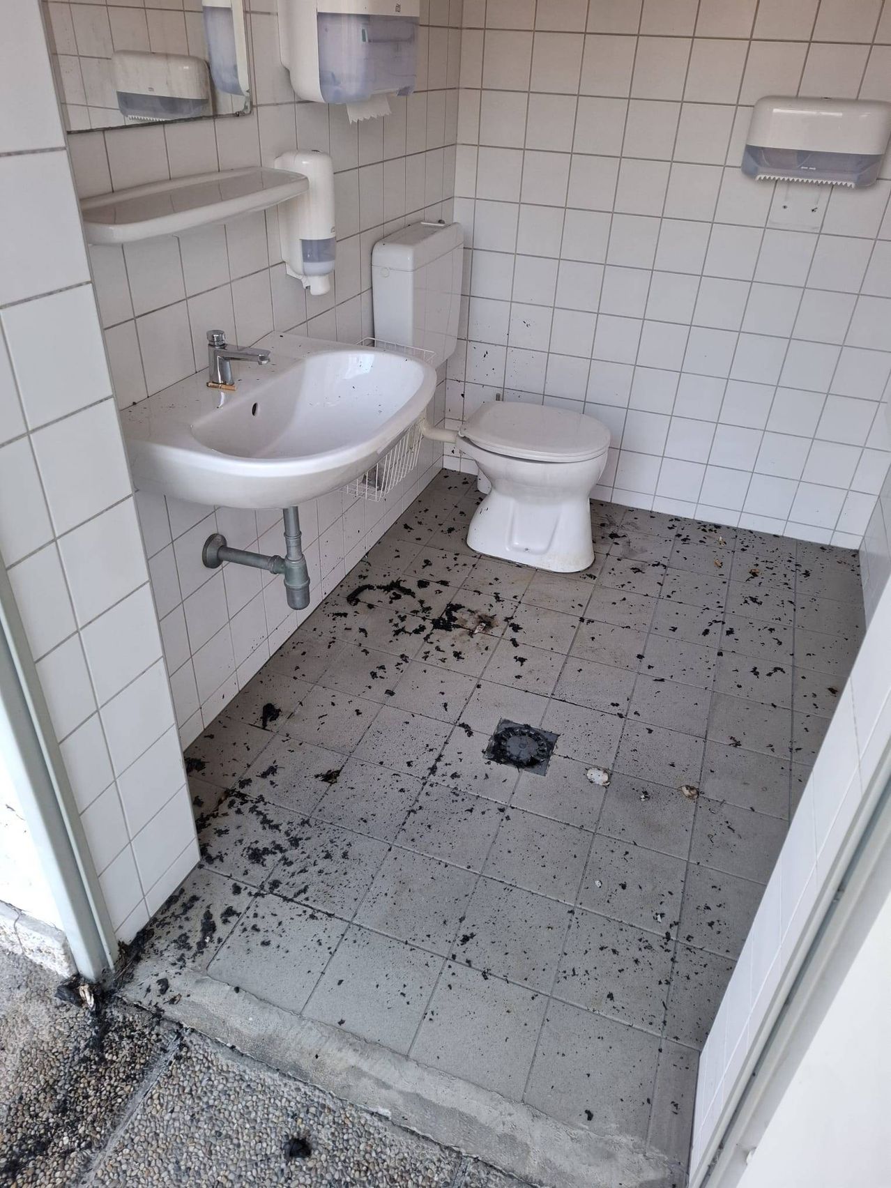 Ein weißes Fliesenbad mit einem Waschbecken und einer Toilette, beide mit schwarzen Rückständen bespritzt, und einem mit schwarzen Rückständen bedeckten Boden.