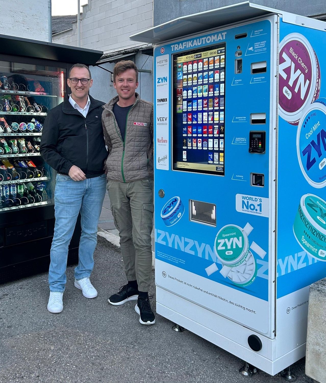 Zwei Männer stehen neben einem blauen Verkaufsautomaten mit verschiedenen Produkten. Ein Mann trägt eine Brille, und der andere hat Turnschuhe an.