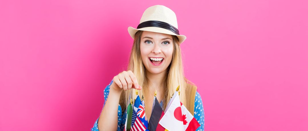 Eine lächelnde Frau mit Hut hält Flaggen verschiedener Länder, einschließlich USA, Australien, Deutschland und Kanada, vor einem rosa Hintergrund.