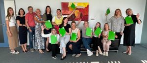 A group of women and men are posing for a photo holding green folders in an office with a colorful painting on the wall.