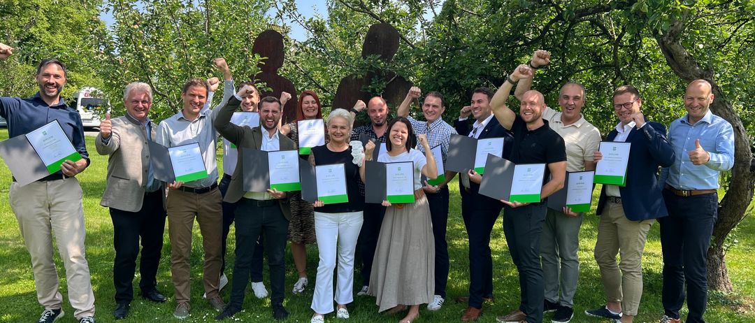 Eine Gruppe von Erwachsenen, sowohl Männer als auch Frauen, stehen in einem Garten zusammen und halten Urkunden in der Hand. Sie lächeln und posieren für ein Foto.