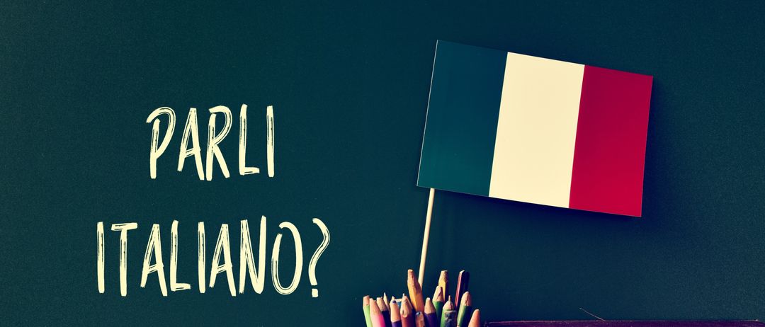 Eine Tafel zeigt den Satz 'Parli Italiano?' neben einer französischen Flagge und einem Stiftehalter. Bücher sind neben der Tafel auf einer Holzoberfläche angeordnet.