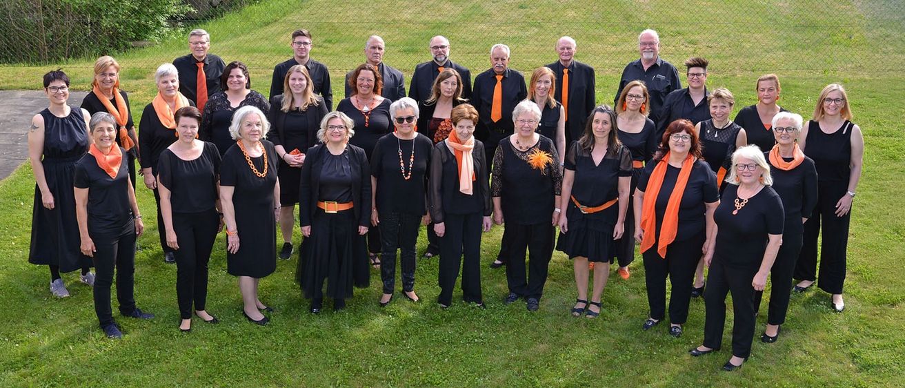 Eine Gruppe von Menschen steht auf einem Rasenfeld, trägt schwarze Kleider und orange Schals. Sie alle lächeln für ein Gruppenfoto.