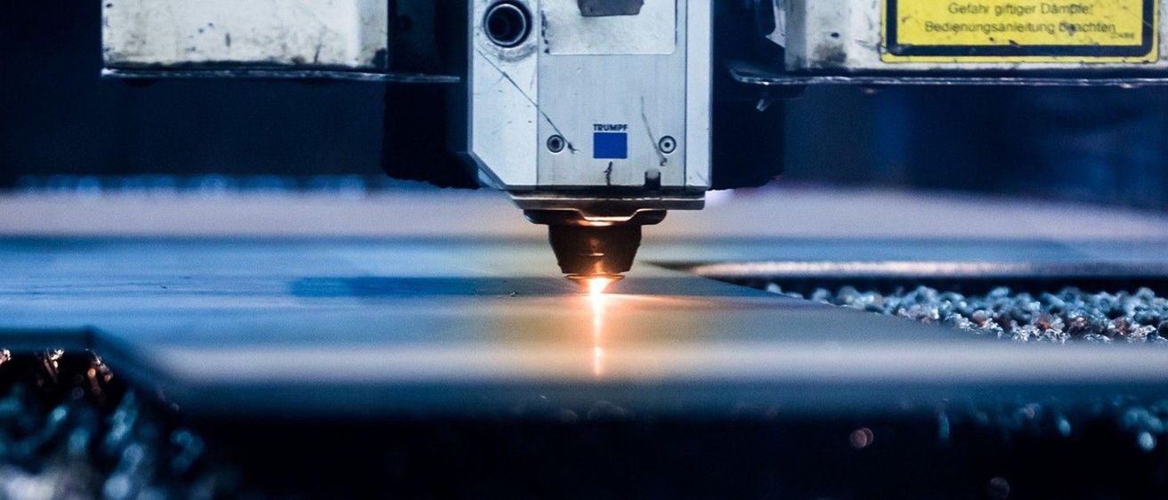 Nahaufnahme einer TRUMPF-Laserschneidmaschine, die eine Metallplatte schneidet. Die Maschine hat einen hellen Lichtstrahl, der auf das Metall fokussiert ist.