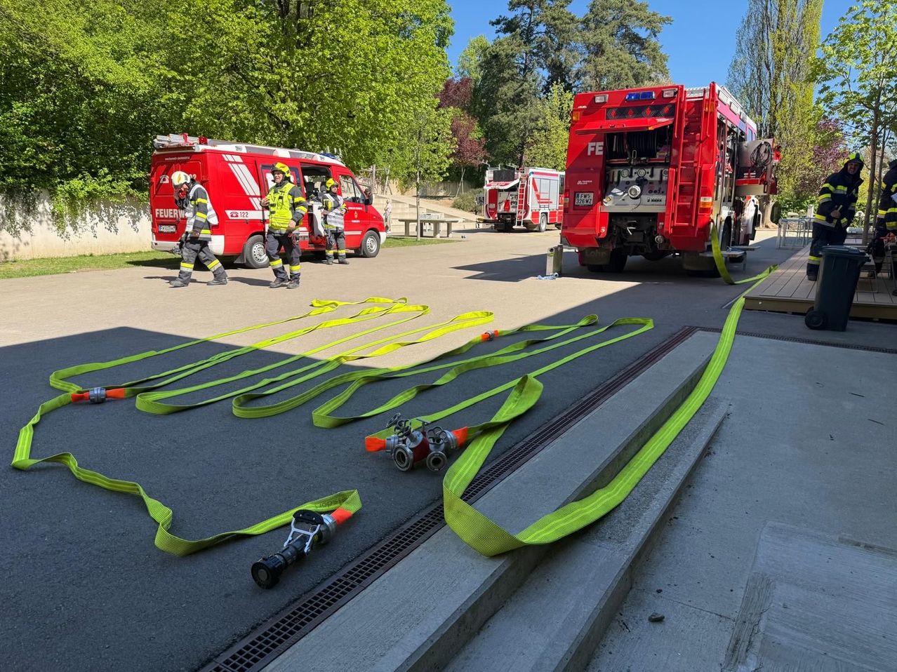 Feuerwehrleute bereiten Ausrüstung an einem sonnigen Tag vor, mit zwei roten Feuerwehrwagen am Straßenrand. Grüne Schläuche liegen auf dem Betonboden aus.