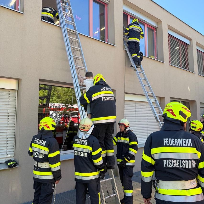Feuerwehrleute klettern eine Leiter an einem Gebäude hoch, tragen Helme und gelb-schwarze Uniformen. Sie retten jemanden aus einem Fenster.