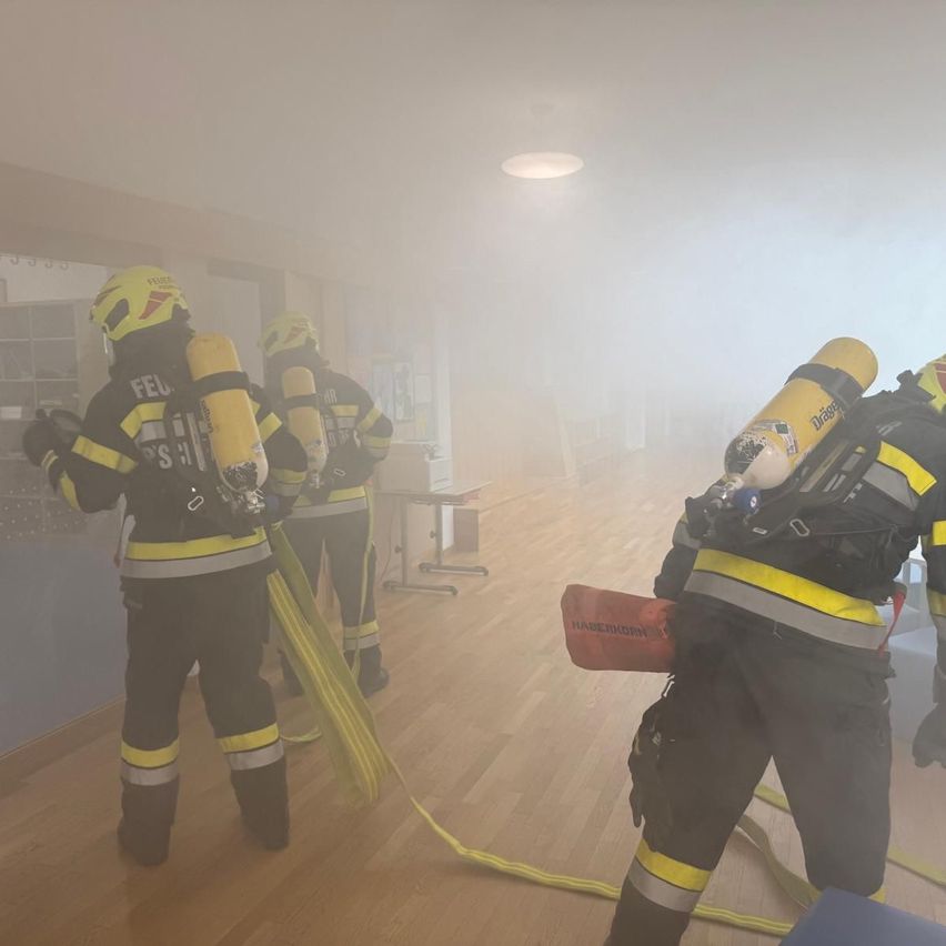 Drei Feuerwehrleute in Schutzkleidung stehen in einem rauchigen Raum und halten Feuerwehrschläuche und Atemschutzgeräte. Sie sind von einem Schreibtisch und einem Schrank umgeben.