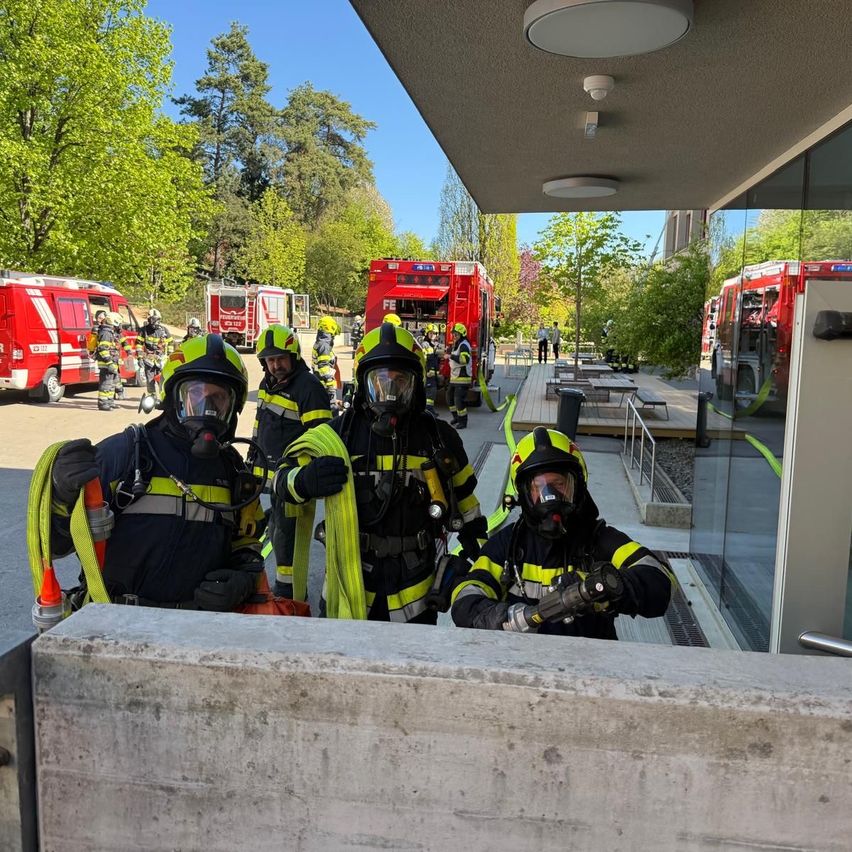 Feuerwehrleute in voller Ausrüstung, einschließlich Masken und Handschuhen, stehen auf einer Plattform. Hinter ihnen sind Feuerwehrwagen auf der Straße geparkt, mit Bäumen und Gebäuden im Hintergrund.