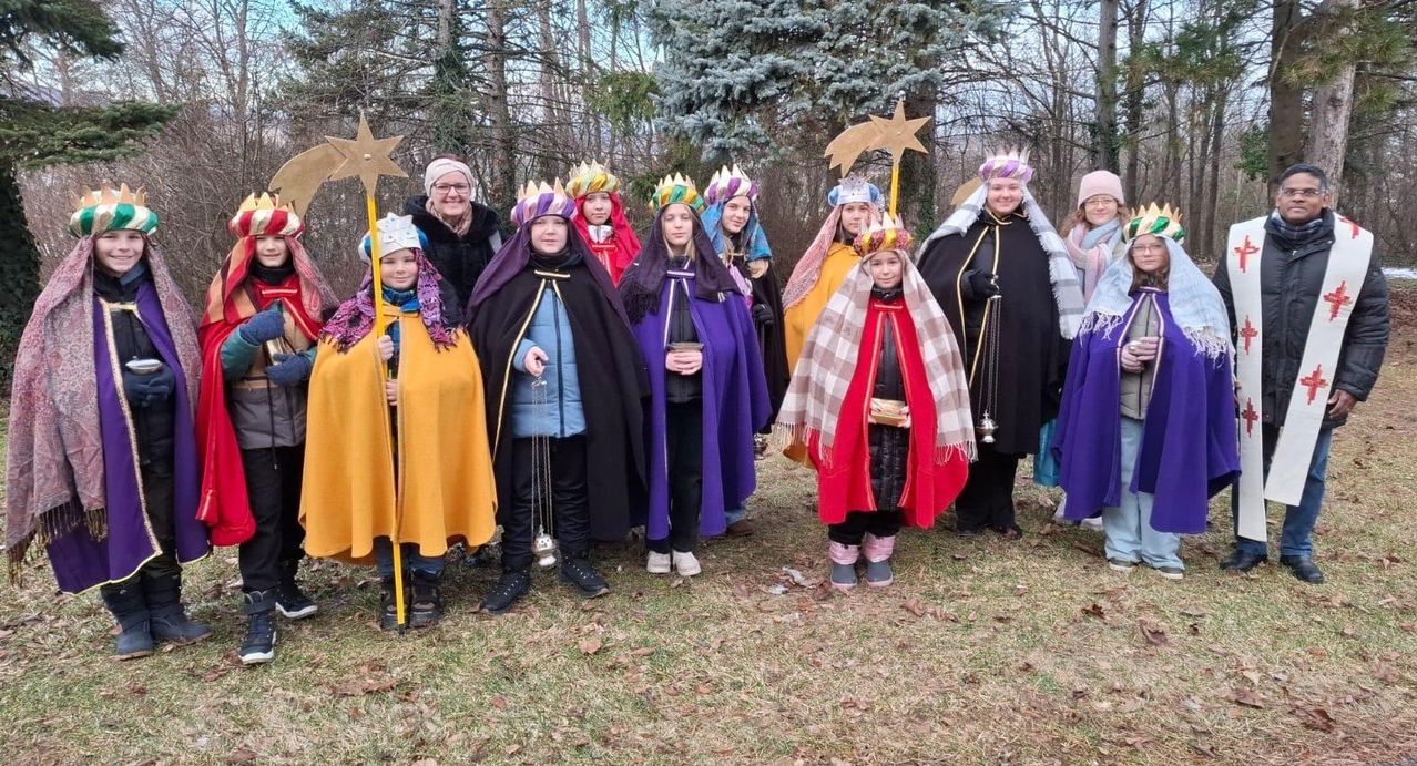 Eine Gruppe von Kindern, die als Könige und Königinnen verkleidet sind, steht in einer Reihe, einige halten sternförmige Requisiten. Sie sind draußen in einem verschneiten Bereich mit Bäumen im Hintergrund.