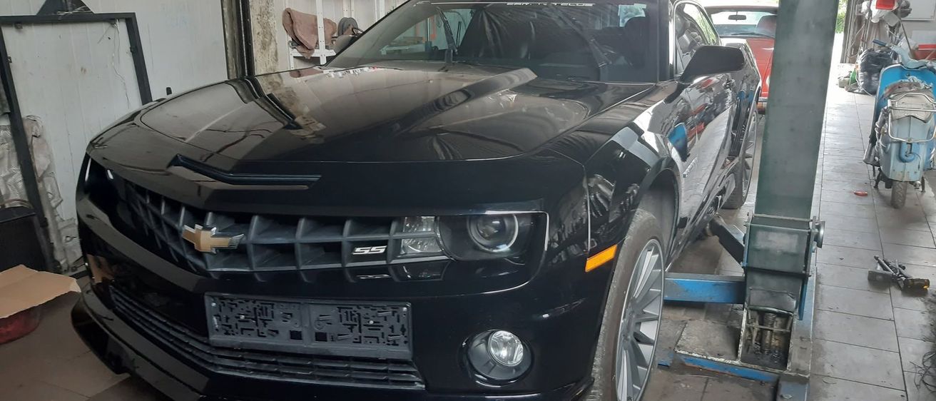 Ein schwarzer Chevrolet Camaro SS steht in einer Garage, wobei die Front des Autos zur Kamera zeigt. Das Auto befindet sich auf einer Autohebebühne, wobei die Vorderräder vom Boden abgehoben sind.