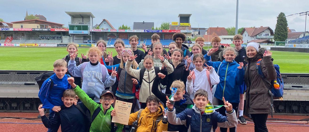 Eine Gruppe junger Athleten posiert auf einer Laufbahn für ein Foto, hält Urkunden und Medaillen in den Händen. Einige lächeln und machen Friedenszeichen. Dahinter sind Tribünen, Häuser und eine Straßenlaterne.