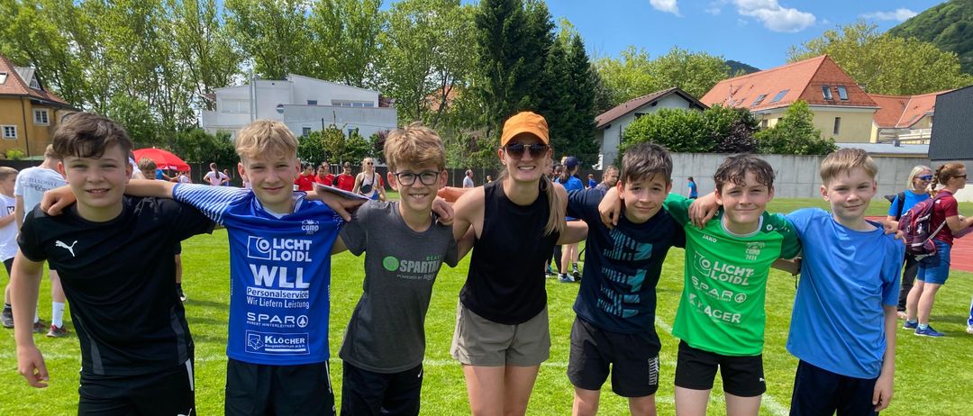Sechs junge Jungen und eine Frau mit oranger Kappe posieren für ein Foto auf einem Rasenfeld, mit Gebäuden und Bäumen im Hintergrund.