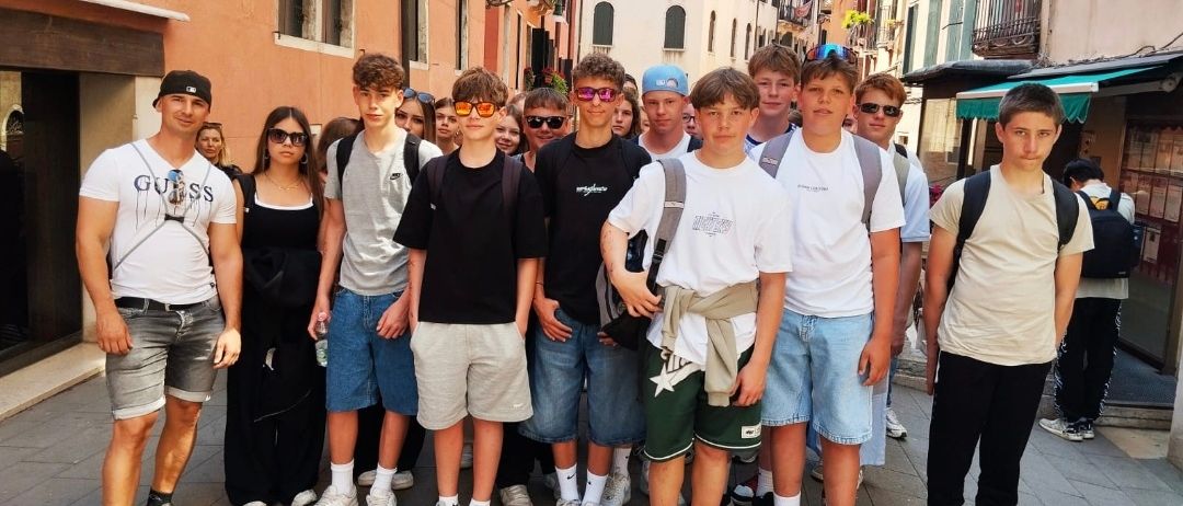 Eine Gruppe junger Männer und Frauen steht zusammen auf einem Bürgersteig in einer Stadt. Sie tragen Freizeitkleidung und einige haben eine Brille. Sie lächeln und scheinen für ein Foto zu posieren. Hinter ihnen befinden sich Gebäude mit Balkonen und Vordächern.
