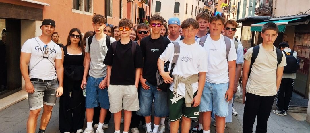 Eine Gruppe junger Männer und Frauen steht zusammen auf einem Bürgersteig in einer Stadt. Sie tragen Freizeitkleidung und einige haben eine Brille. Sie lächeln und scheinen für ein Foto zu posieren. Hinter ihnen befinden sich Gebäude mit Balkonen und Vordächern.