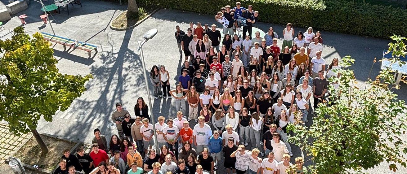 Eine große Gruppe von Menschen steht in einem Innenhof, umgeben von Bäumen und Büschen. Im Hintergrund sind Gebäude zu sehen, eines mit einem schwarzen Dach. Tische, Bänke und ein Spielplatz sind ebenfalls vorhanden.