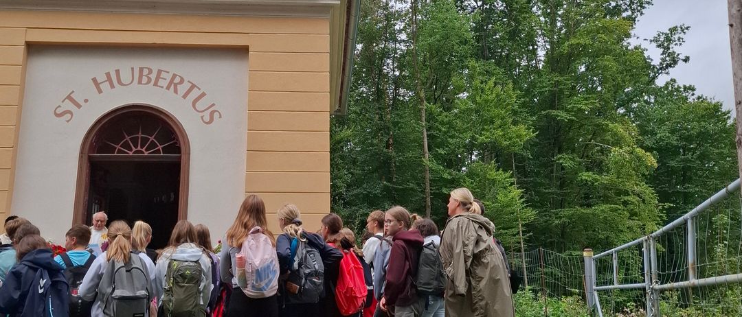 Eine Gruppe von Schülern steht vor der Kapelle St. Hubertus, einige tragen Rucksäcke und Turnschuhe. Die Kapelle ist gelb mit einer roten Kuppel, und ein Metallzaun ist in der Nähe.