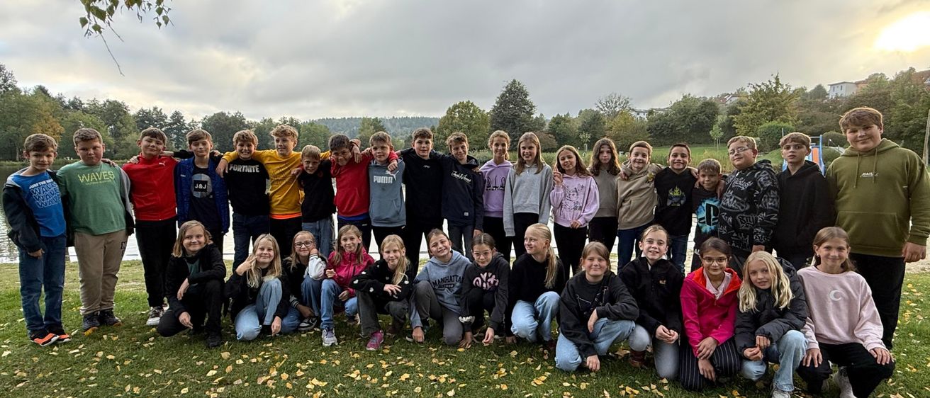 Eine Gruppe junger Menschen posiert für ein Foto in einem Grasbereich mit Bäumen im Hintergrund. Sie stehen und sitzen auf dem Gras.