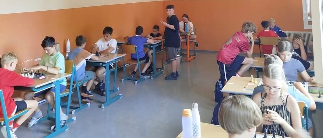 In einem Klassenzimmer sind mehrere Kinder in einer Schachpartie engagiert. Der Raum hat Tische, Stühle und Schachfiguren. Die Wände sind in einem orange-weißen Farbschema gestrichen.