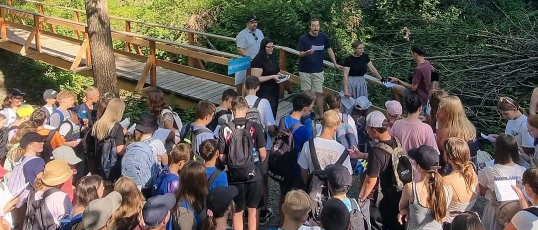 Eine Gruppe von Menschen steht in einem Wald, einige tragen Rucksäcke. Zwei Männer stehen auf einer Holzbrücke, einer hält ein Klemmbrett. In der Nähe unterhalten sich mehrere Personen. Die Umgebung ist üppig grün.