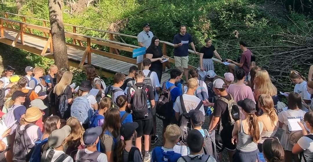 Eine Gruppe von Menschen steht in einem Wald, einige tragen Rucksäcke. Zwei Männer stehen auf einer Holzbrücke, einer hält ein Klemmbrett. In der Nähe unterhalten sich mehrere Personen. Die Umgebung ist üppig grün.