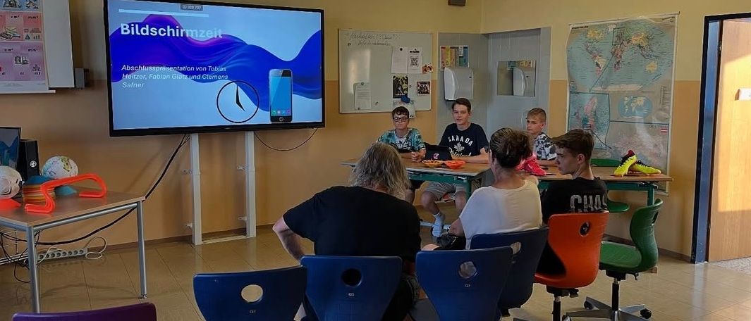 Gruppe von Schülern sitzt um einen Tisch in einem Klassenzimmer herum, auf einem Monitor ist Bildschirmzeit zu sehen. Poster und eine Karte schmücken die Wände.