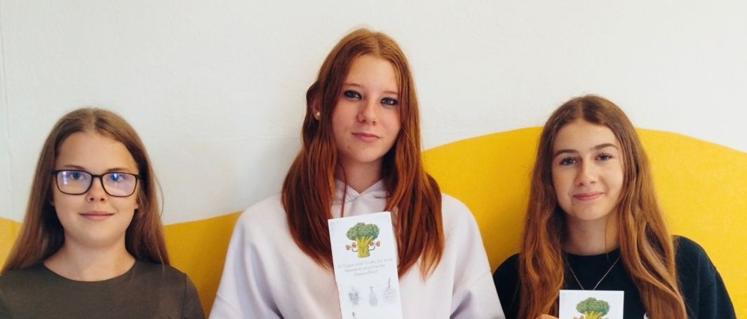 Drei junge Frauen stehen nebeneinander und halten ein Papier in der Hand. Sie lächeln und posieren für ein Foto. Die Frau auf der linken Seite trägt eine Brille. Die Frau in der Mitte trägt eine weiße Sweatshirt und hält ein Papier mit einem Brokkoli-Zeichnung. Die Frau auf der rechten Seite trägt ein schwarzes Langarmshirt.