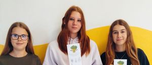Drei junge Frauen stehen nebeneinander und halten ein Papier in der Hand. Sie lächeln und posieren für ein Foto. Die Frau auf der linken Seite trägt eine Brille. Die Frau in der Mitte trägt eine weiße Sweatshirt und hält ein Papier mit einem Brokkoli-Zeichnung. Die Frau auf der rechten Seite trägt ein schwarzes Langarmshirt.