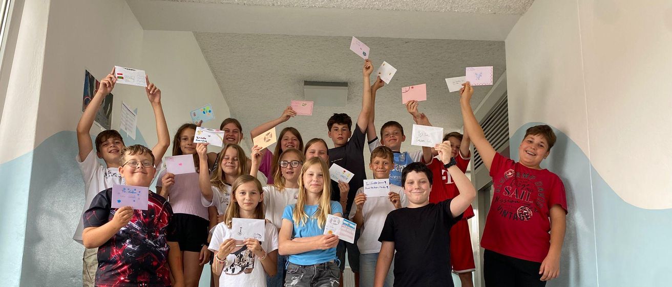 Eine Gruppe von Kindern steht auf den Stufen, alle halten kleine Zettel in der Hand, und einige heben ihre Hände. Sie alle lächeln und schauen zur Kamera.