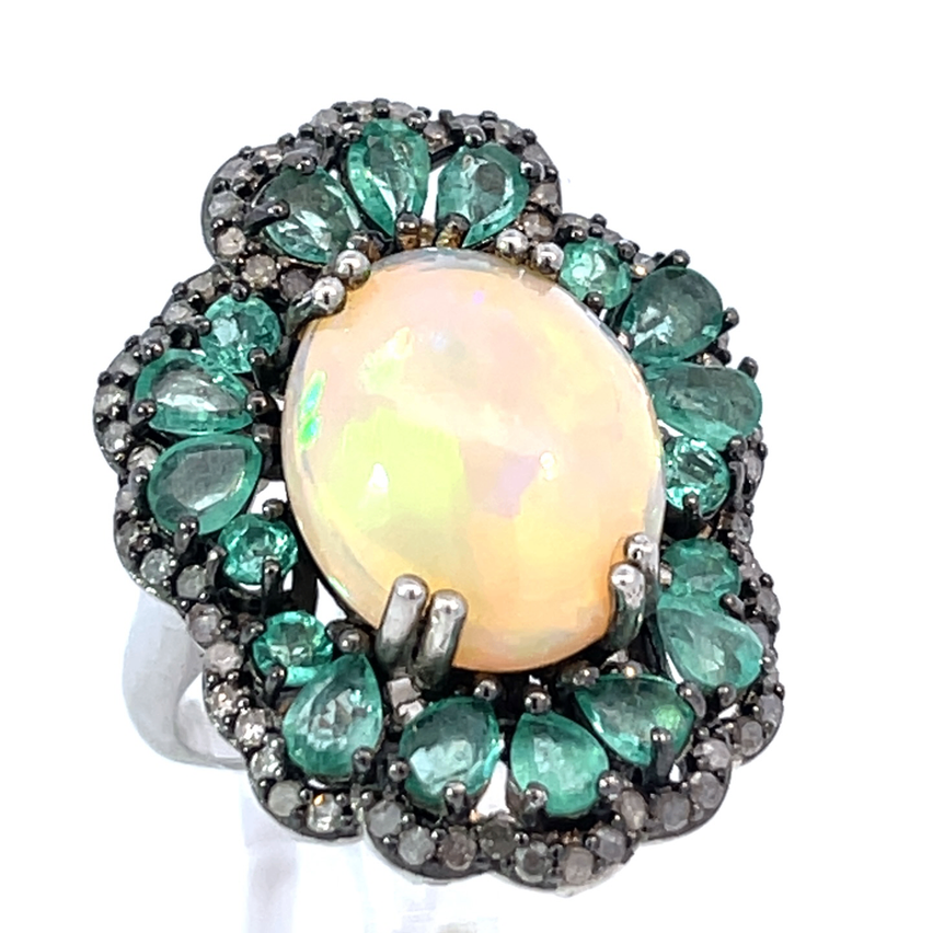 Ein Silberring hat einen ovalen Opal-Zentralstein mit einem gelben Farbton, umgeben von grünen Edelsteinen in Smaragdform. Das Setting umfasst verschiedene graue und schwarze Steine.