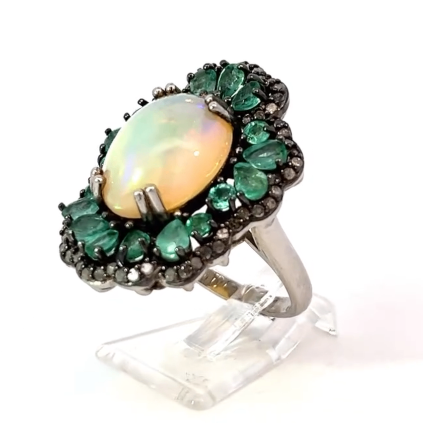 Ein Silberring mit einem großen ovalen Opal-Zentralstein, umgeben von grünen Smaragden und schwarzen Diamanten, präsentiert auf einem klaren Ständer.