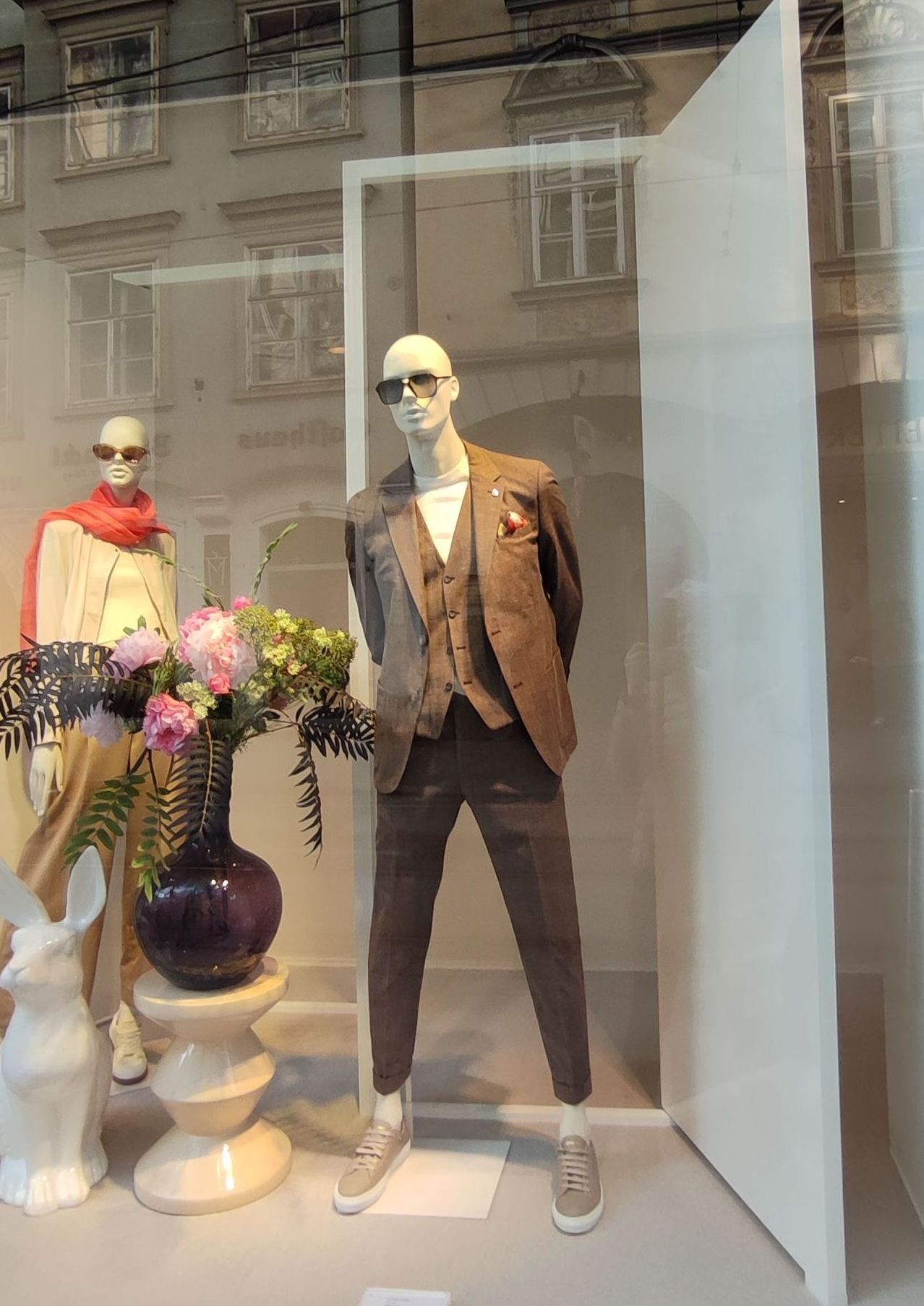 Ein Dummy in einem braunen Anzug und Weste steht vor einer Glasvitrine, neben einer Blumenvase und einer Kaninchenstatue.