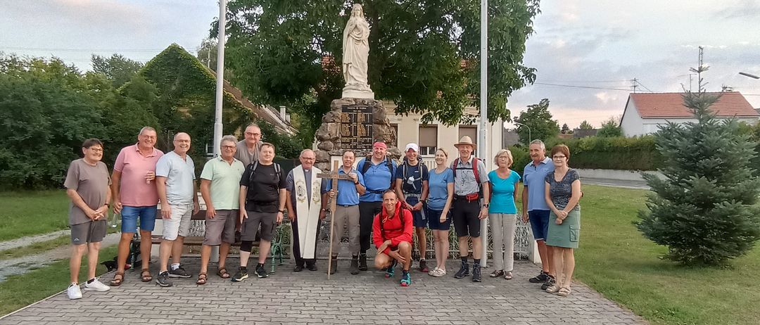 Eine Gruppe von Menschen in Wanderausrüstung, einige mit Hüten, posiert für ein Foto vor einer Statue der Jungfrau Maria.