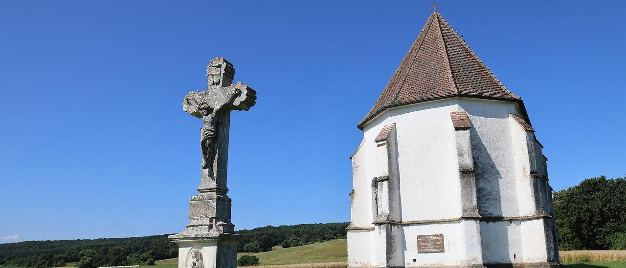 Ein Steinkreuz steht vor einer kleinen Kapelle, beide umgeben von einem Grasfeld unter einem klaren blauen Himmel.