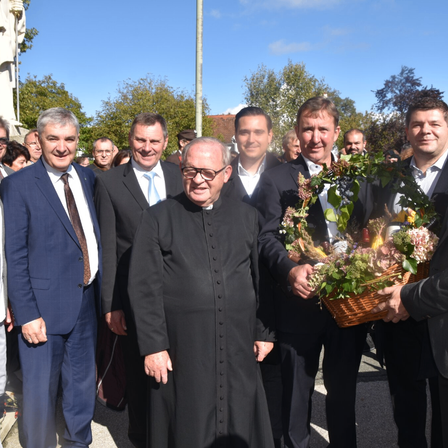 Eine Gruppe von Männern in formeller Kleidung steht im Freien. Ein Mann in einem schwarzen Gewand hält einen Korb mit Blumen. Die anderen lächeln.