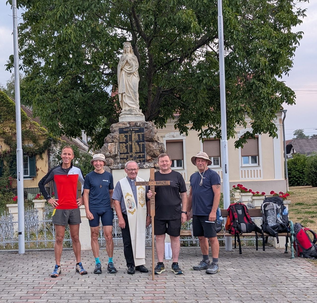 Fünf Männer stehen vor einer Statue und einem Gebäude, mit Rucksäcken und einem Kreuz sichtbar. Ein Mann hält ein Kreuz, und sie tragen alle Freizeitkleidung.