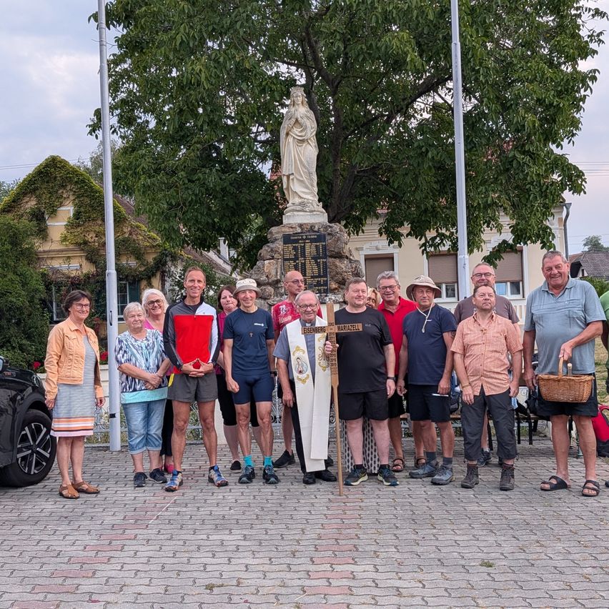 Eine Gruppe von Menschen steht vor einer Statue, einige halten ein Holzkreuz und einen Korb, mit einem Gebäude und Bäumen im Hintergrund.