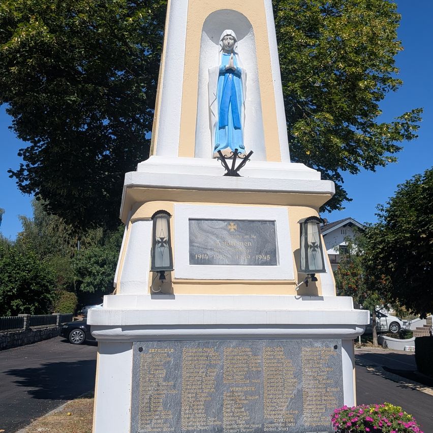 Ein hoher weißer Denkmal mit einer Statue einer Frau in Blau oben. Unter der Statue befindet sich eine Tafel mit Text und Daten. Das Denkmal ist von Grün umgeben und hat eine Tafel mit Namen und Text auf der Vorderseite.