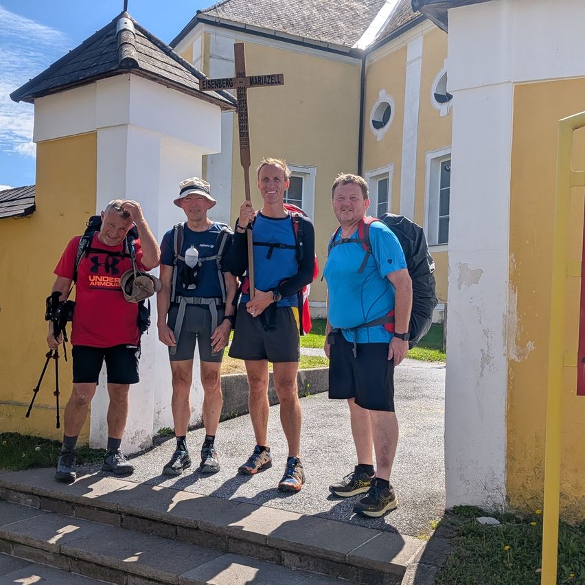 Vier Männer in Wanderausrüstung stehen vor einem Gebäude mit einem Schild, das Eisenberg und Mariazell anzeigt.