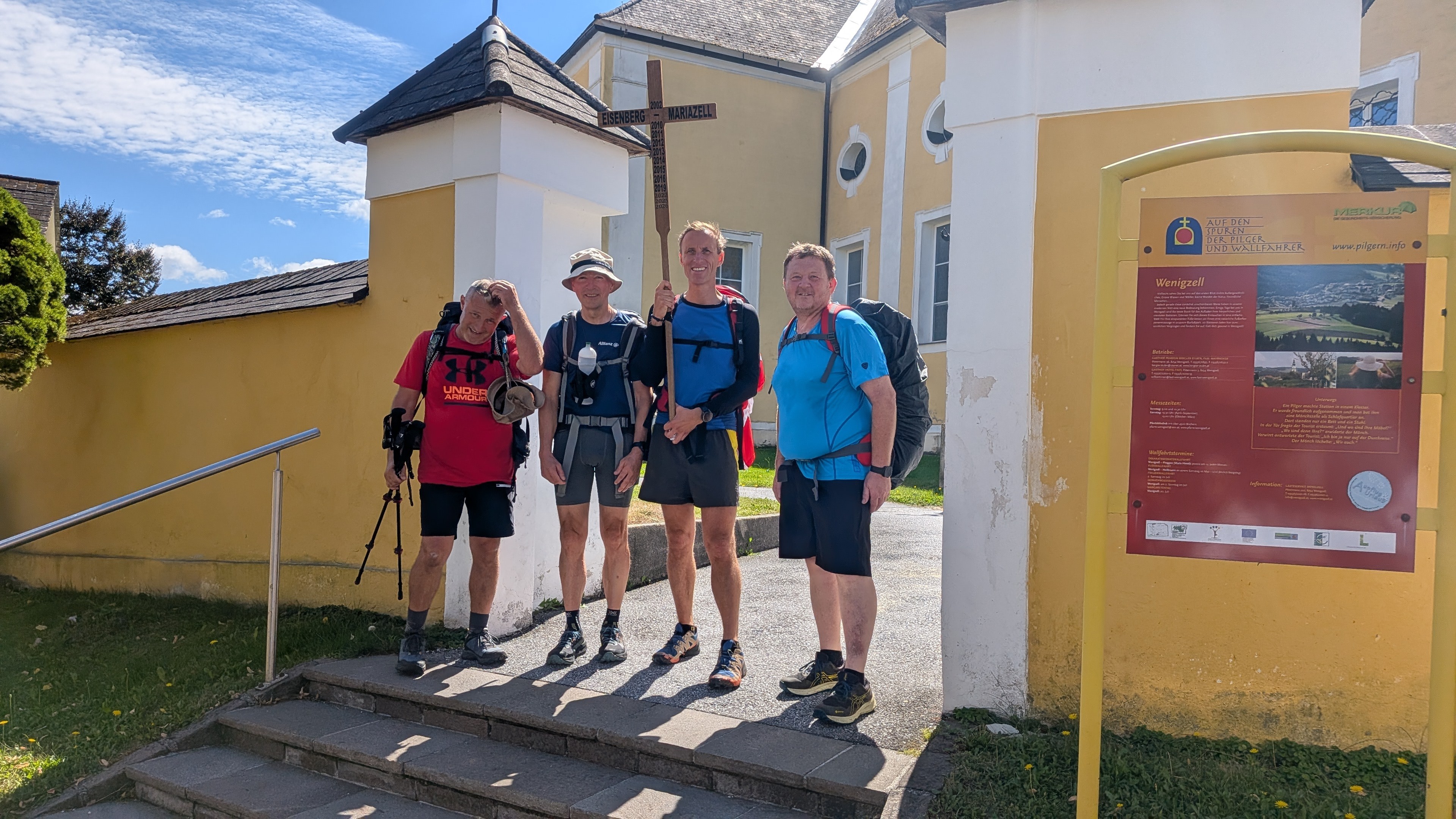 Vier Pilger mit Rucksäcken vor einem gelben Gebäude mit Kreuzweg-Schild in Wenigzell