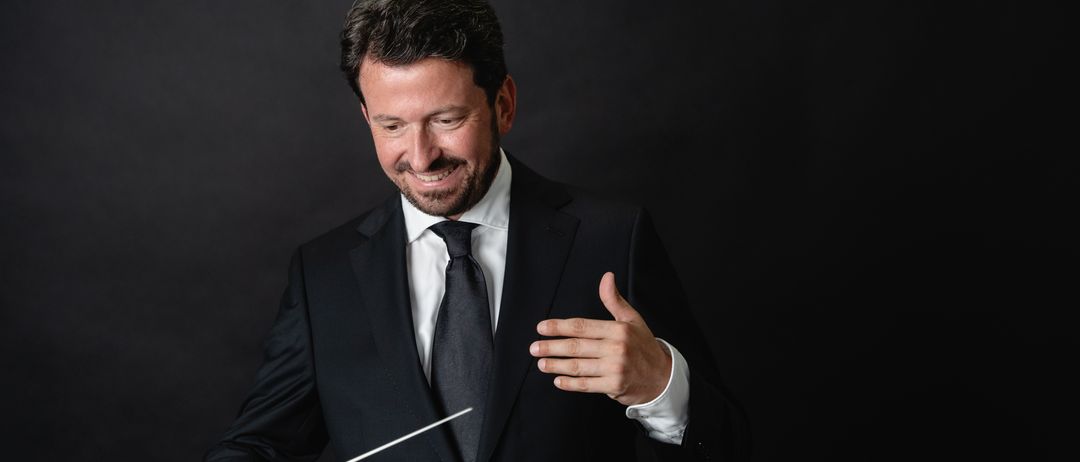 Ein Mann in einem schwarzen Anzug und Krawatte, lächelnd, gestikuliert mit einem Taktstock, wahrscheinlich dirigiert er.