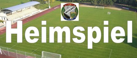 Eine Draufsicht auf ein Fußballfeld mit dem Text 'Heimspiel' in weißer Schrift. Das Feld ist leer, und es gibt ein Logo in der Mitte. Das Feld ist von einem Zaun umgeben, und es gibt Bäume und ein Gebäude in der Ferne.