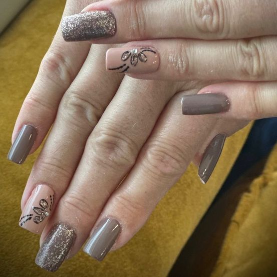 Ein Nahaufnahme der Hand einer Person mit fünf gepflegten Fingernägeln. Jeder Nagel ist in einer anderen Grau- und Silbernuance lackiert. Zwei Nägel haben Blumendesigns mit silbernem Glitzer. Der Hintergrund ist gelb.