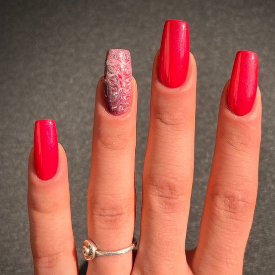 Eine Hand mit knallroten Nägeln, mit einem einzigartigen Marmordesign auf dem Mittelfinger und einem silbernen Ring am Ringfinger.