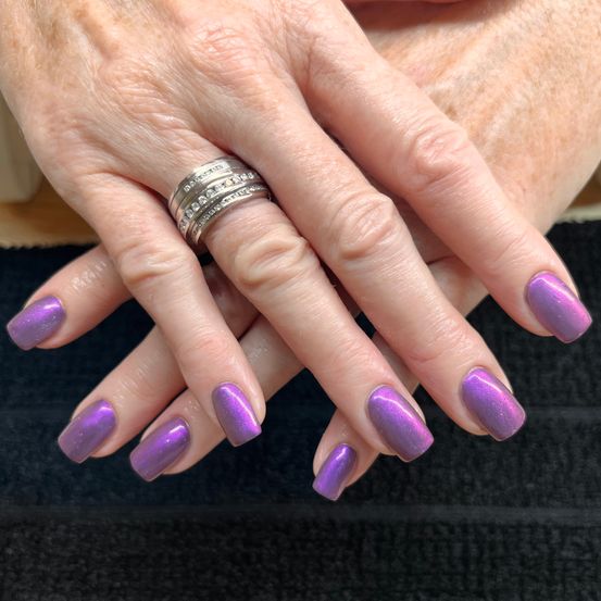 Die Hände einer Person mit gepflegten Nägeln, die in einem glänzenden Lila-Ton lackiert sind. Eine Hand trägt einen silbernen Ring mit Edelsteinen.