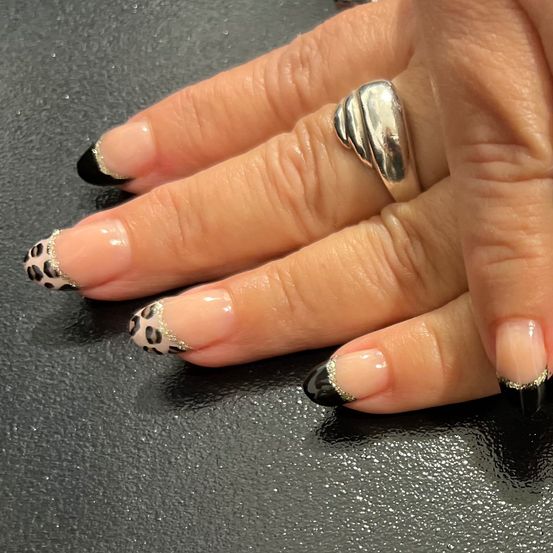 Ein Nahaufnahme der Hand einer Person mit Leopardenmuster in Schwarz-Weiß lackierten Nägeln, drei silbernen Ringen und einer schwarzen Oberfläche.