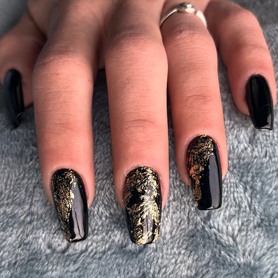 Ein Nahaufnahme einer Hand mit schwarzem und goldenem Nagellack auf einem grauen Pelzteppich. Die Nägel haben einen glänzenden, glitzernden Effekt.