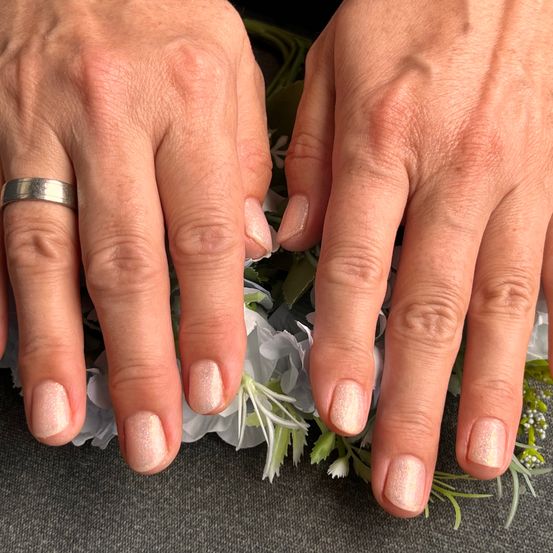 Zwei Hände mit gepflegten Nägeln ruhen auf einem Bett aus weißen Blumen. Die linke Hand trägt einen Ring. Die Nägel sind in einer hellen, schimmernden Farbe lackiert.