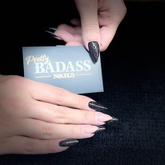 Eine Person hält eine Visitenkarte mit der Aufschrift 'Pretty Badass Nails' und schwarzem Nagellack. Die Karte ist weiß mit goldenem Text.