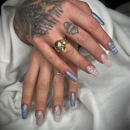 Eine Hand mit detailliertem Tattoo und einem goldenen Totenkopfring zeigt glitzernden blauen und rosa Nagellack auf einem weißen Tuch.